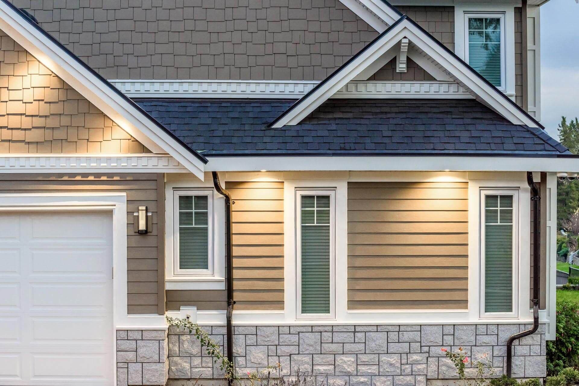 Modern house exterior with charcoal roof, stone foundation, horizontal siding, white trim, three windows, garage door on left, and landscape lighting
