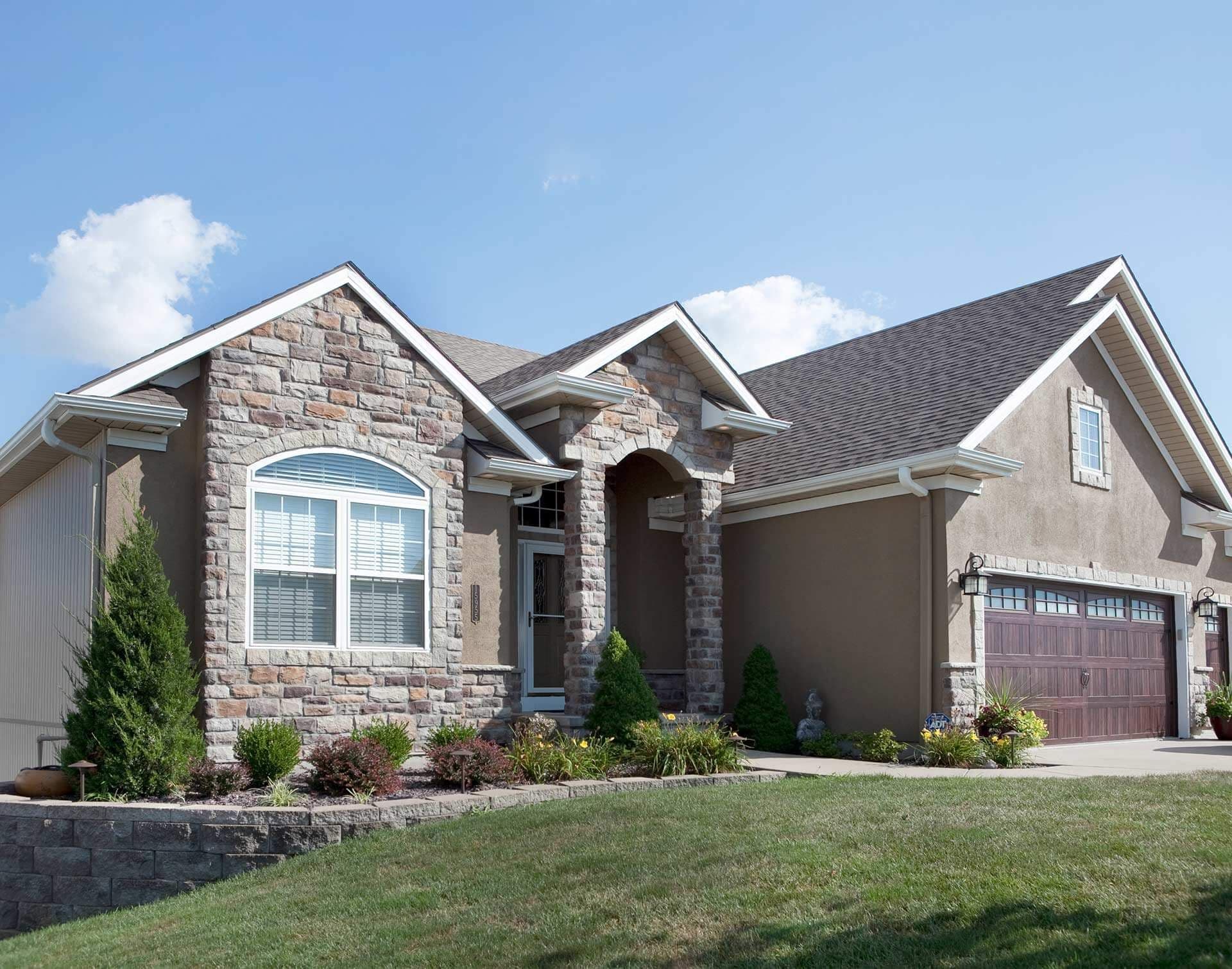 Single-story residential home with stone and stucco exterior, arched entryway, two-car garage, and manicured landscaping under blue sky