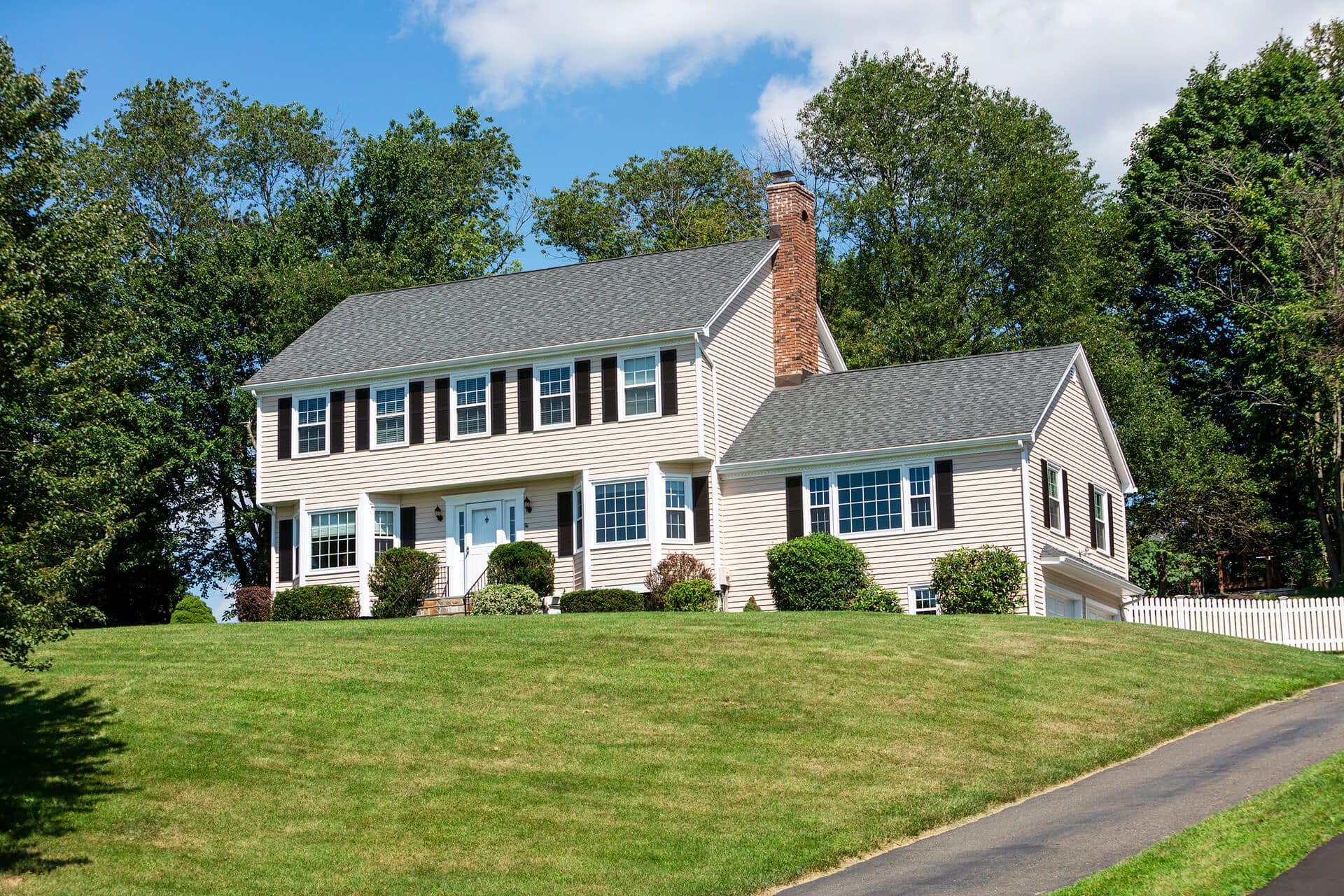 Two-story colonial house with white siding, brick chimney, and black shutters surrounded by landscaped lawn and trees